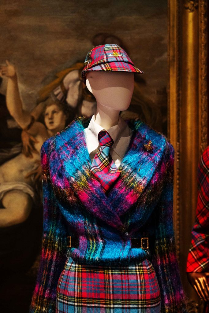 A mannequin dressed in colourful tartan jacket and hat in a red-walled stately home gallery space 