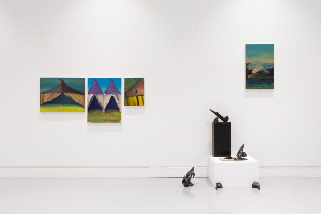 A wall in a gallery. To the left, three paintings, with another higher on the right. On the floor against the wall to the right, a cluster of low plinths and dark ceramic works.