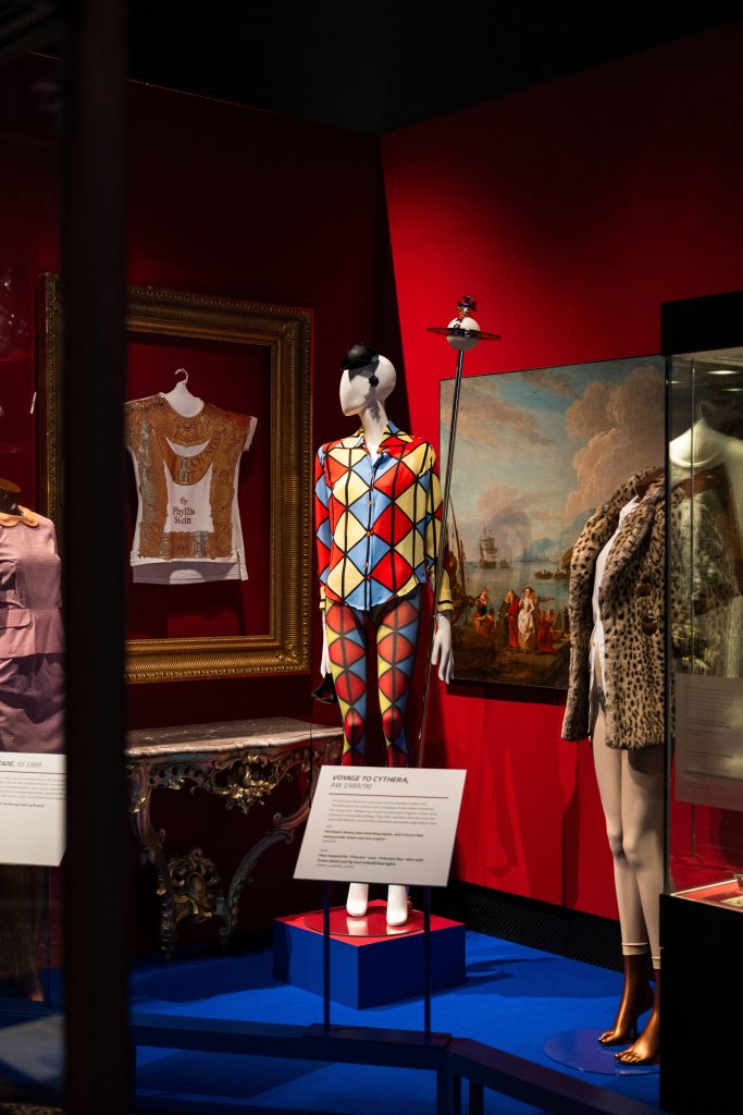 A mannequin dressed in a harlequin shirt and trouser twinset in a red-walled stately home gallery space