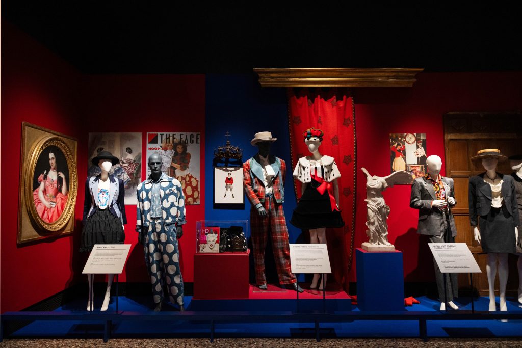 A  number of mannequins dressed in extravagant couture outfits in a red-walled stately home gallery space 
