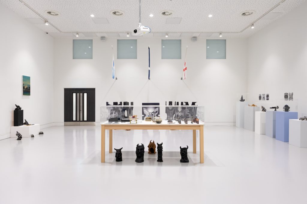 A large whitewall gallery space with scukptural objects with AI imagery against the back wall, a table containing various art objects in the centre, and ceramic objects gathered beneath the table and along the side walls