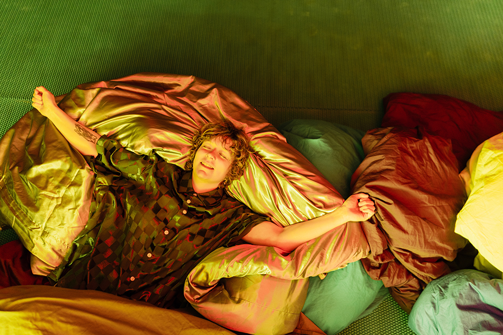 A person lies on a pile of duvets, arms outstretched a look of happy relaxation on their face.