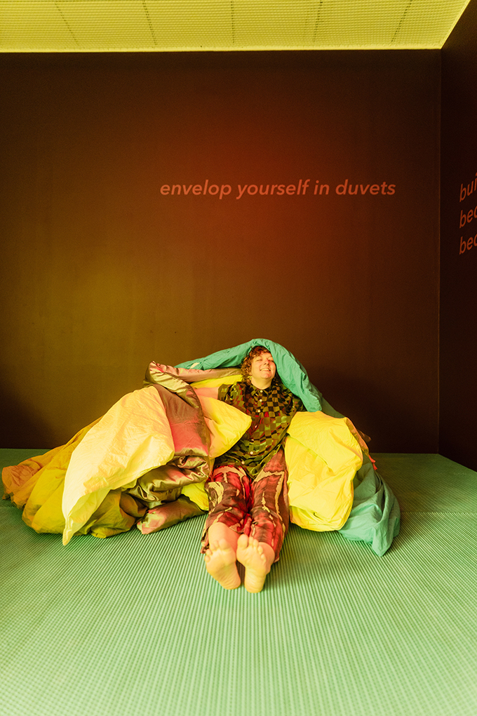 A person wrapped in duvets under a sign that reads, 'envelop yourself in duvets'.
