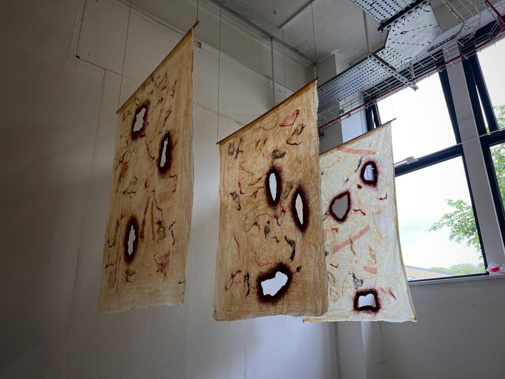 Three large sheets of fabric are suspended from the ceiling, each resembles mammalian skin with holes of rot or burning broken through