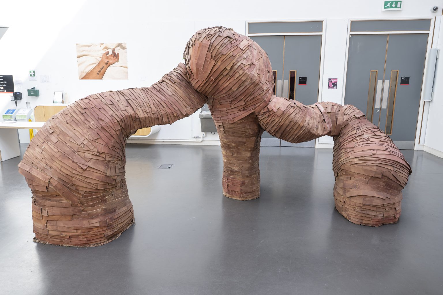 A tentacle like sculpture on three legs made of wound sections of brown card or paper emerges from the floor of an institutional workspace
