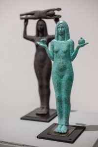 two sculptures of women. each woman is naked with shoulder length hair. one woman holds a child above her head. the other woman holds an apple core in one hand and a pomegranate in the other.