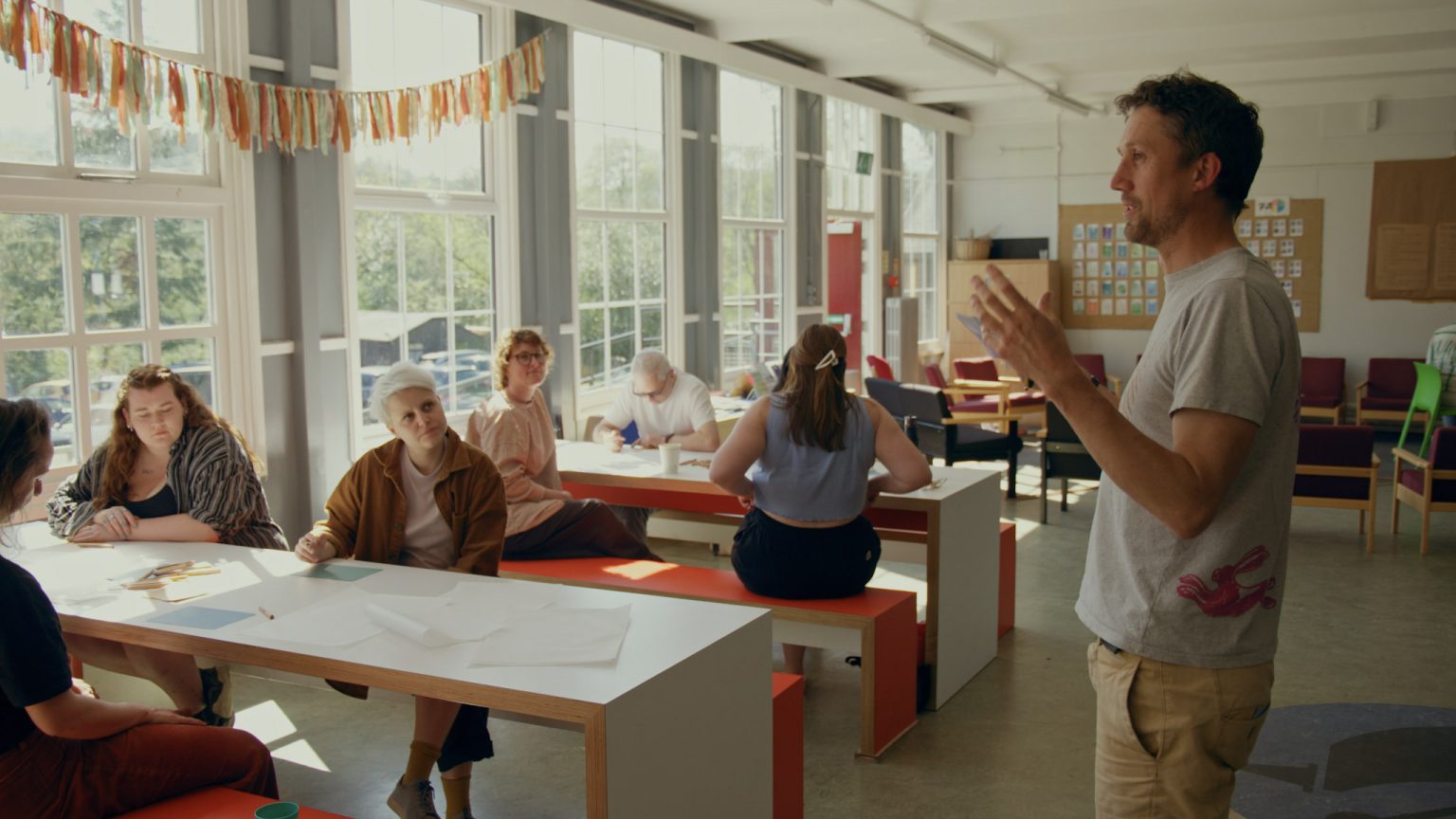 A classroom with bright light coming in the windows and a man at the front addressing the people seated at tables