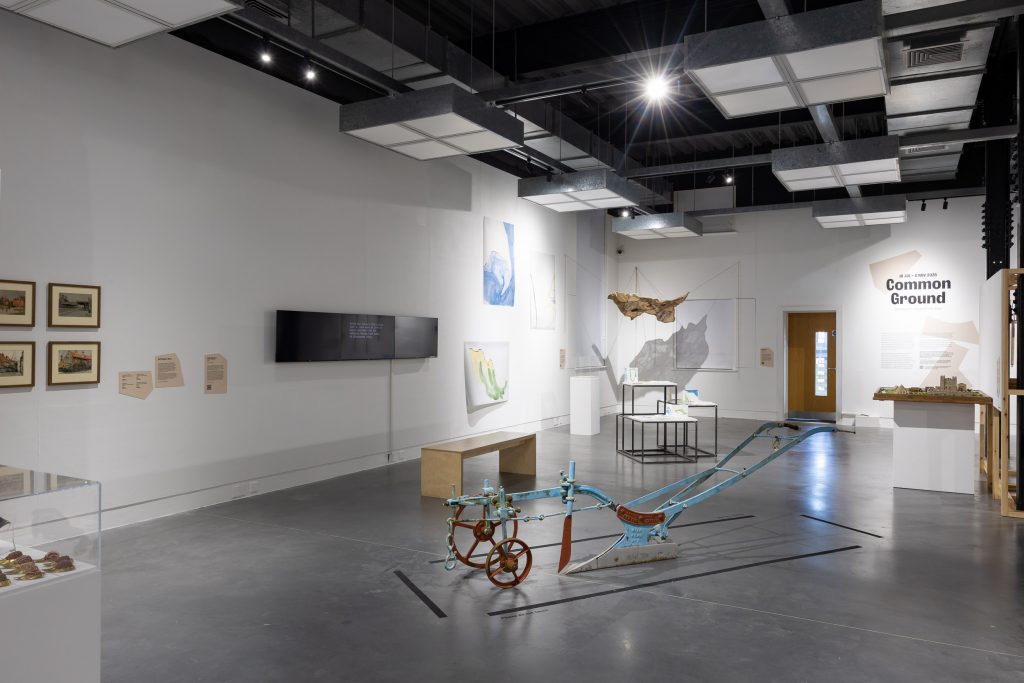 A wide view of the gallery at The Hub with a metal plough in the centre and various different contemporary artworks presented around the walls, including framed photographs, screen-based works and sculptures suspended from the ceiling.