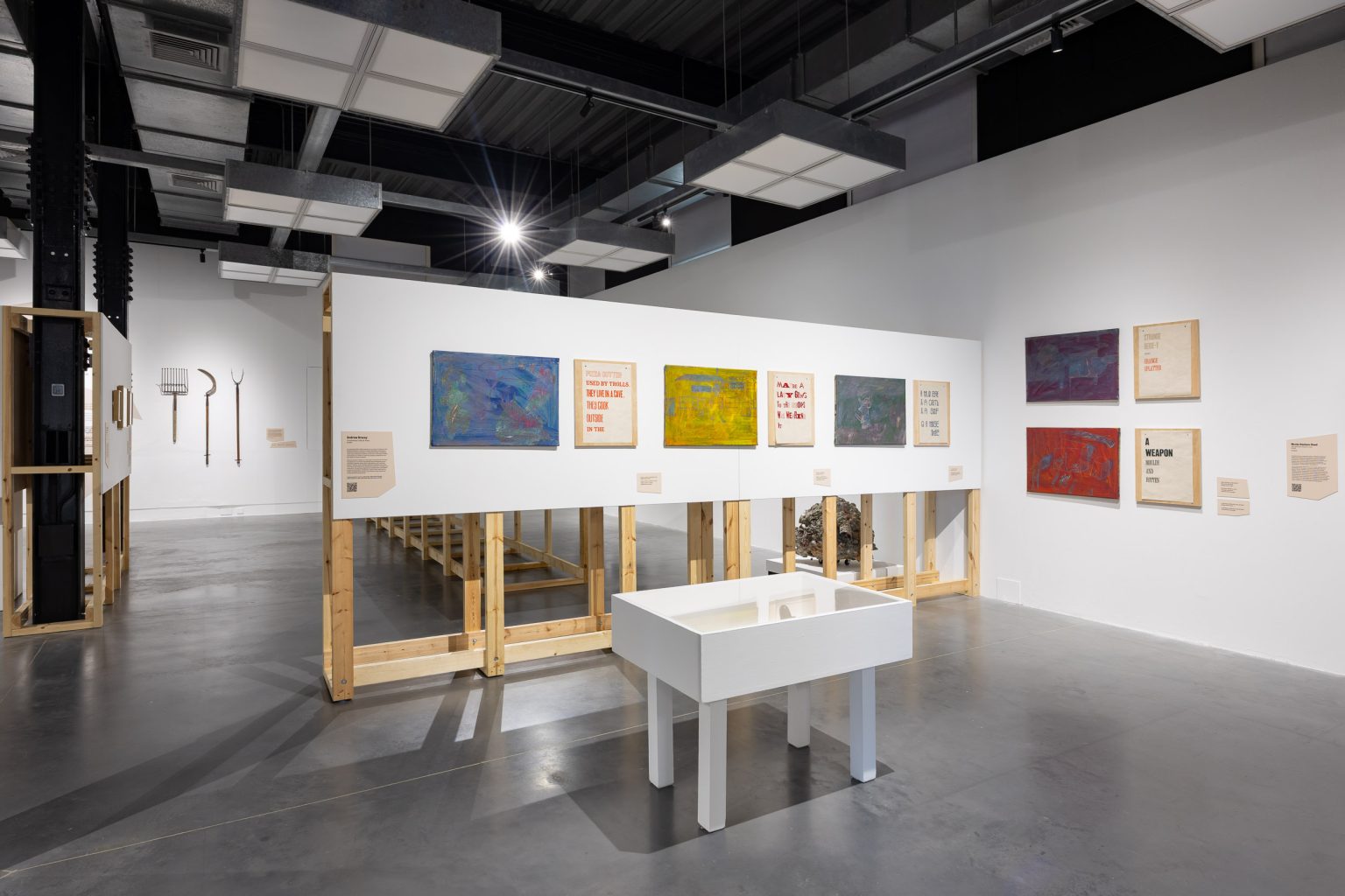 An installation view in a white walled gallery space. Several wall based works are shown around the walls with a small display case in front. The works seem to be a combination of abstract paintings in different primary colours and text-based printed matter.
