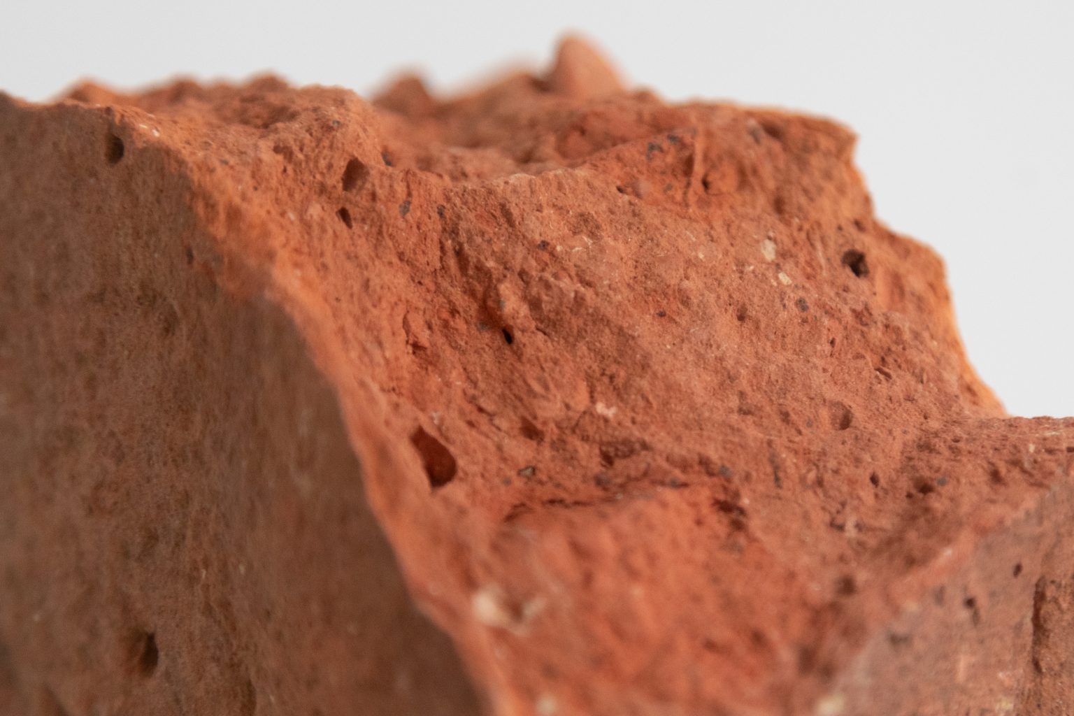 A close up view of a broken red brick, with rough textures, tiny stones and other fragments visible.