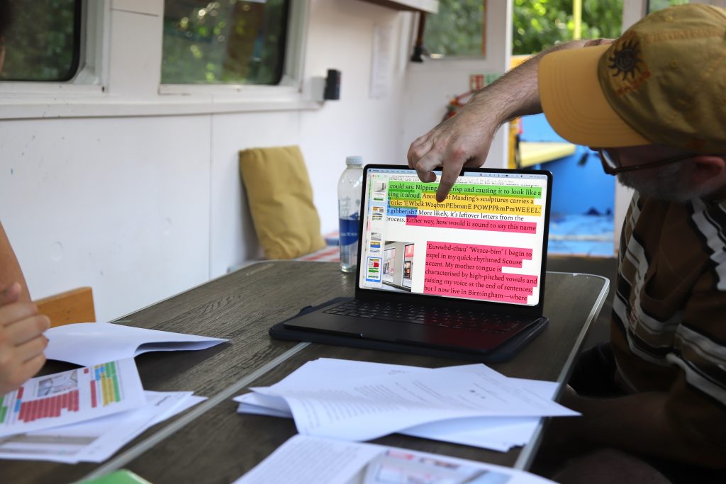 The editor points at his laptop screen, text highlighted in different colours, whilst participants watch and edit their words, all aboard a canal boat.