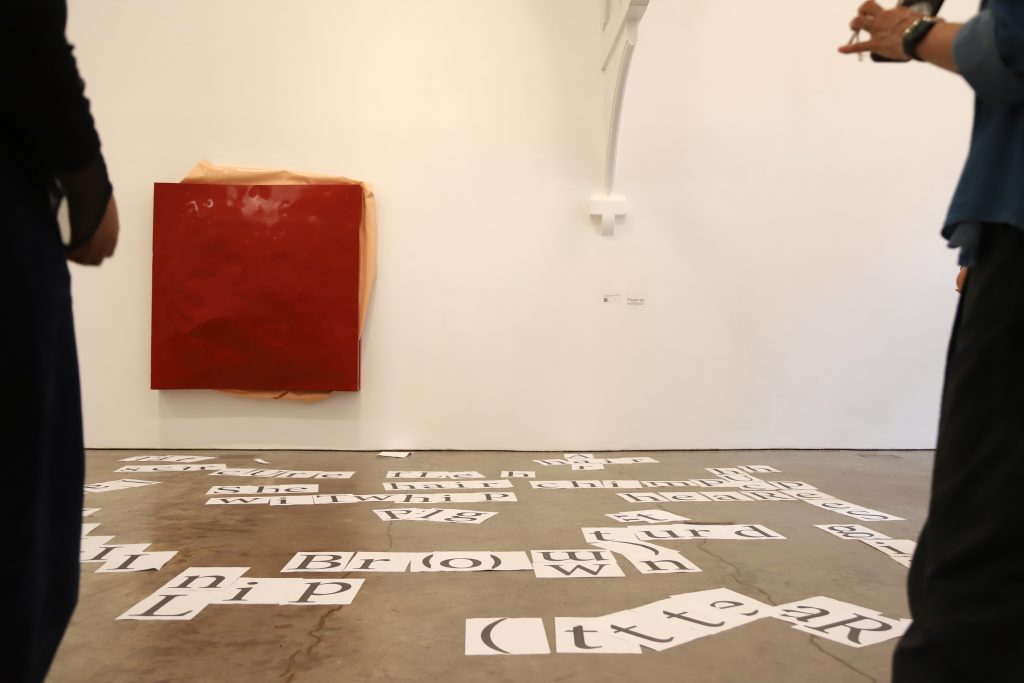 A4 print outs of words and letters are scattered across the gallery floor under a red 'bloated' painting by Angela de la Cruz.