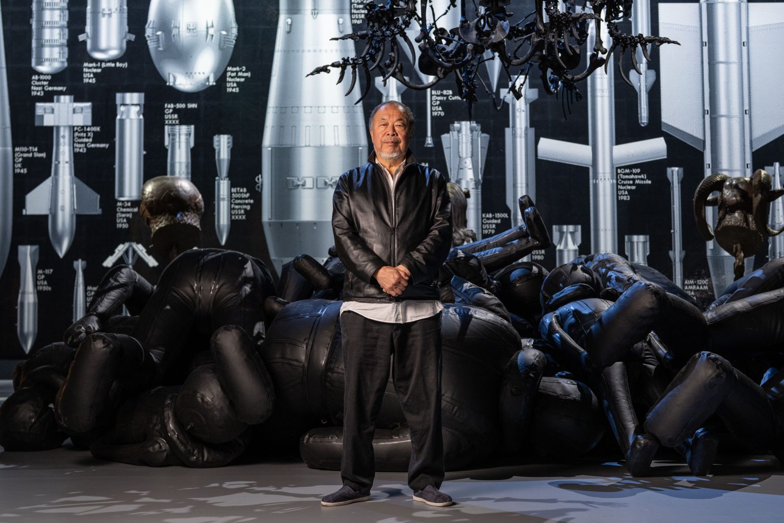 The artist wears stands with hands clasped proudly in front of the black inflatable sculptures of refugees and the wall depicting life sized bombs