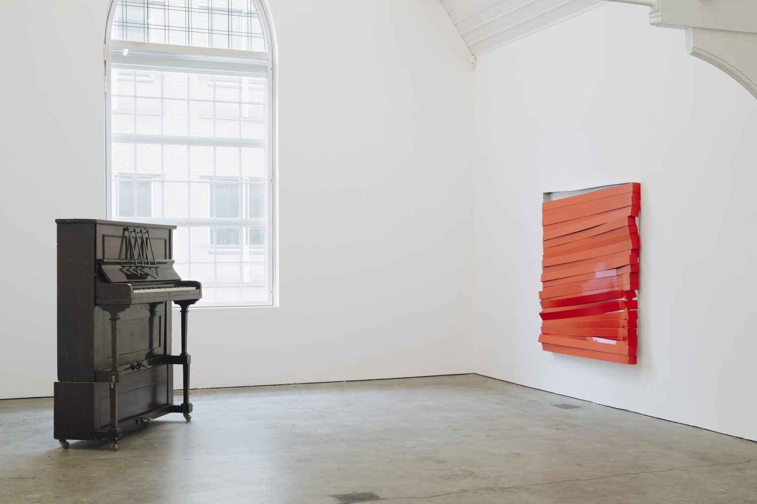 2 artworks by Angela de la Cruz, on the left, a work made from a functioning piano, on the right, a red slatted metal painting is semi-broken, hung on the wall