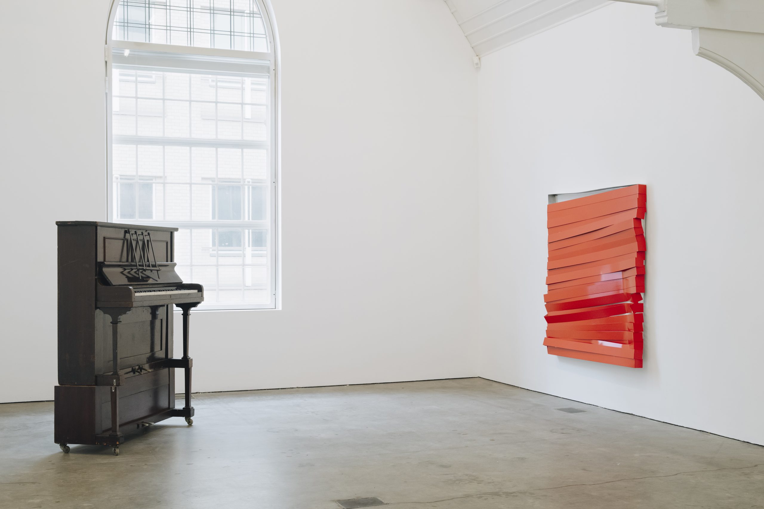 2 artworks by Angela de la Cruz, on the left, a work made from a functioning piano, on the right, a red slatted metal painting is semi-broken, hung on the wall