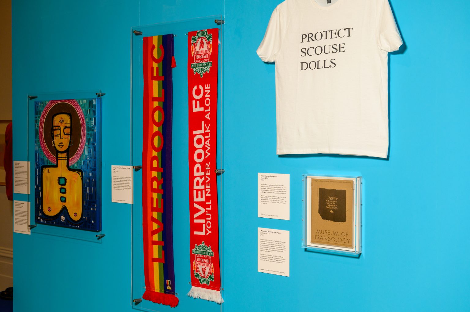 Installation shot showing a collection of objects: A Liverpool football scarf woven in pride colours, a t-shirt with the text 'protect scouse dolls', an abstract image depicting a woman's head and bare chest, and a piece of paper printed with the words 'museum of transology'