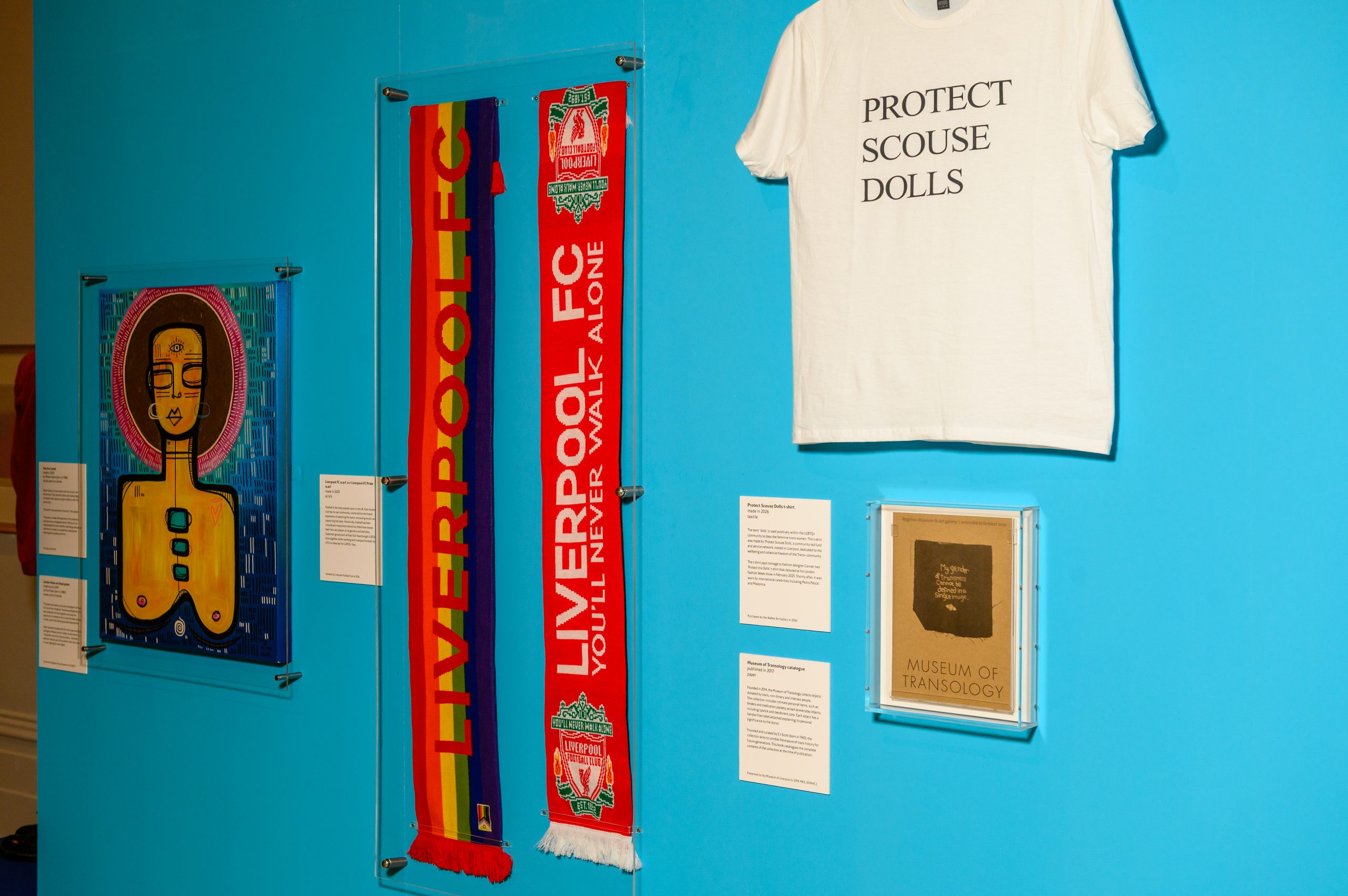 Installation shot showing a collection of objects: A Liverpool football scarf woven in pride colours, a t-shirt with the text 'protect scouse dolls', an abstract image depicting a woman's head and bare chest, and a piece of paper printed with the words 'museum of transology'