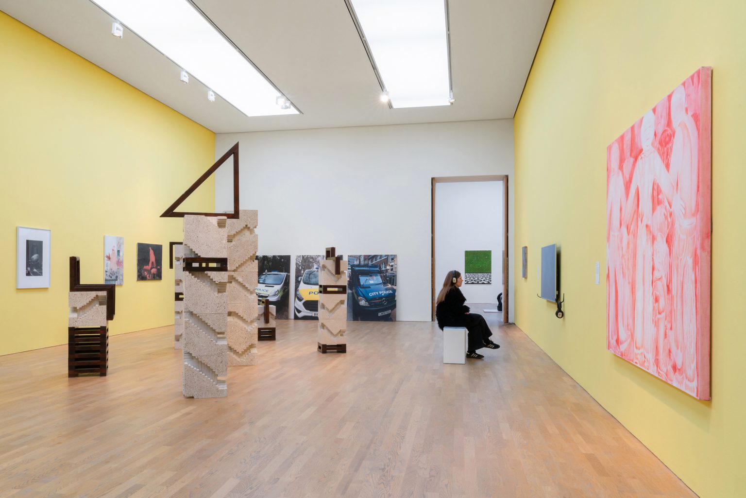 A gallery space with laminated wood floor and yellow painted walls. A pinkish painting on the right, block sculptures in the middle, a person sat on a bench watching a video wearing headphones.