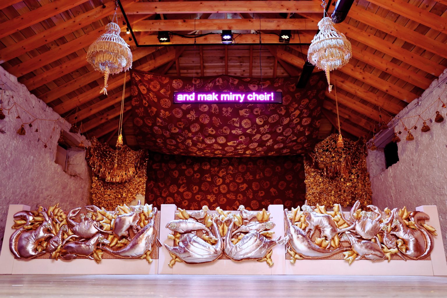 An opulent room with a stage and curtains and chandeliers. An led screen reads 'and mek mirry cheir!'