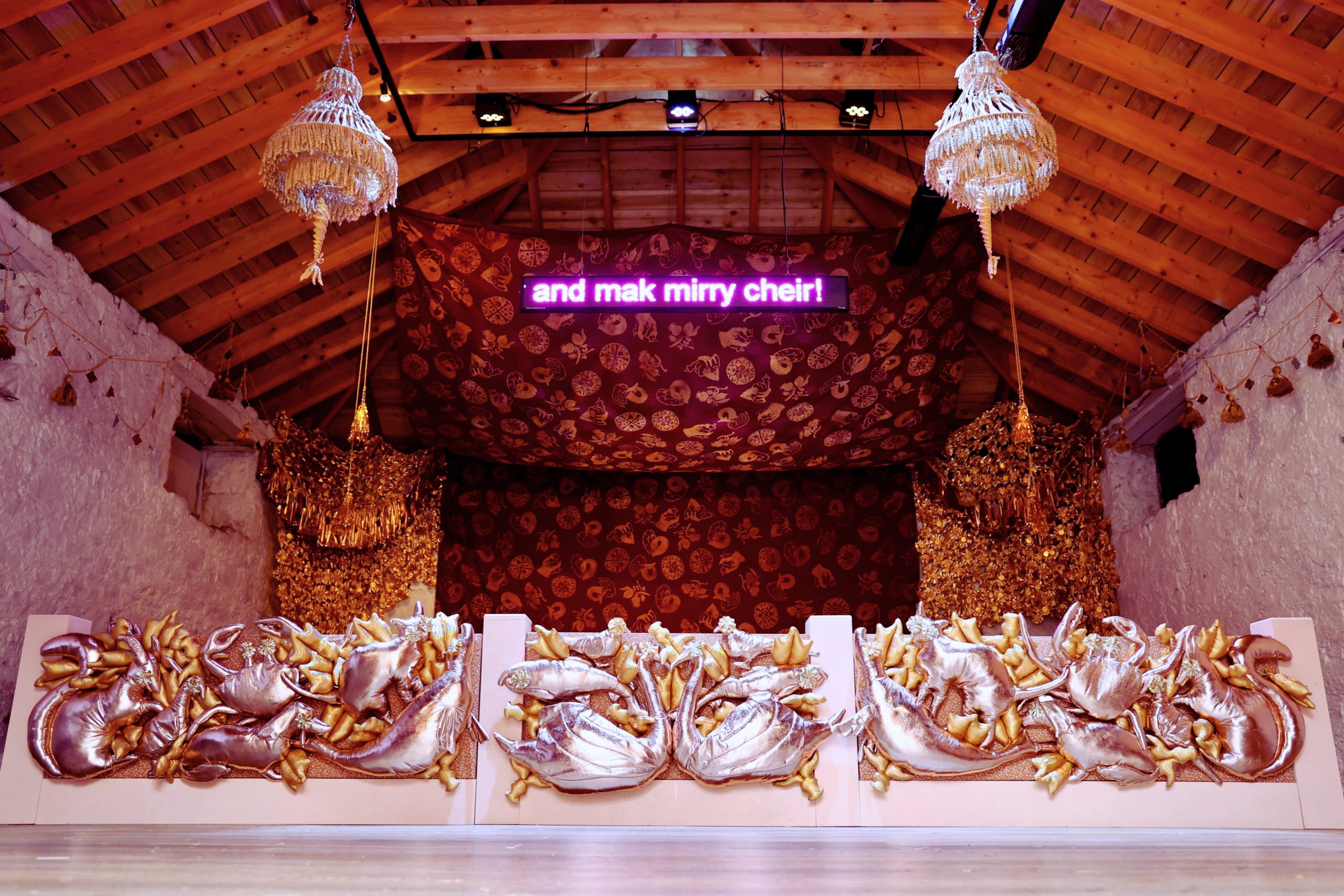 An opulent room with a stage and curtains and chandeliers. An led screen reads 'and mek mirry cheir!'