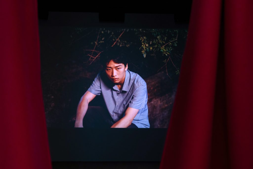 A still from a film, shows a young man in a blue shirt looking thoughtful. On either side and partially obscuring the screen is a red curtain.