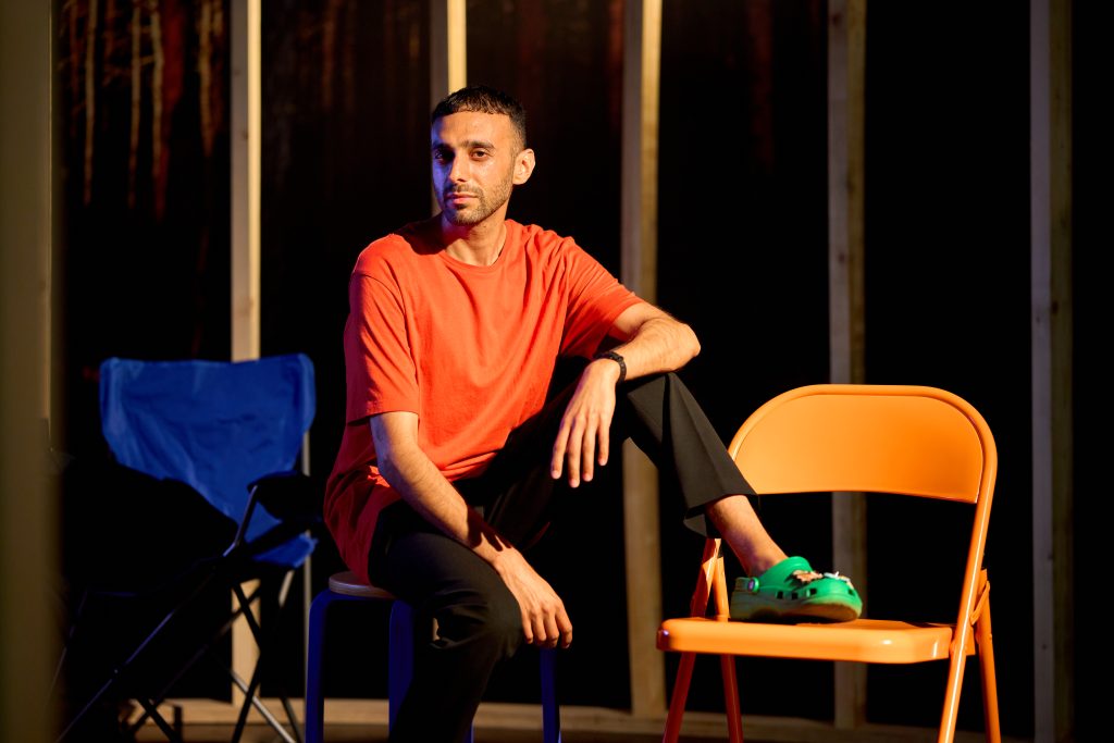 Sahjan Kooner wears an orange T shirt and looks at the camera. One leg is on the floor, the other rests on an orange chair, wearing green Crocs. 