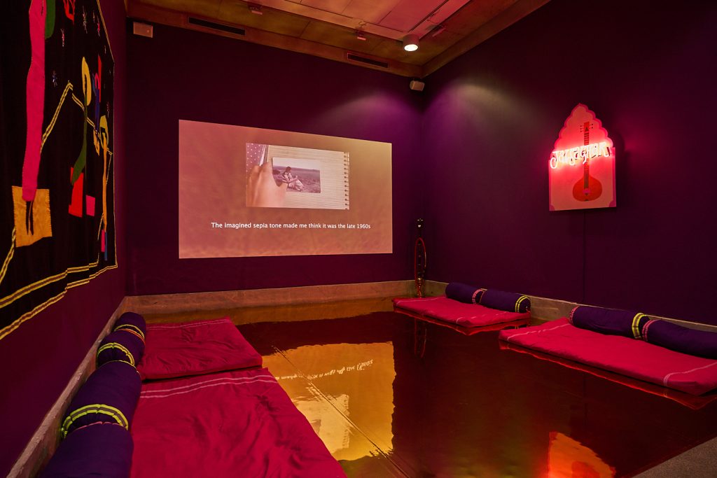 A dark room with no natural light has a golden, mirrored floor and deep purple walls. A film is projected on the far wall.