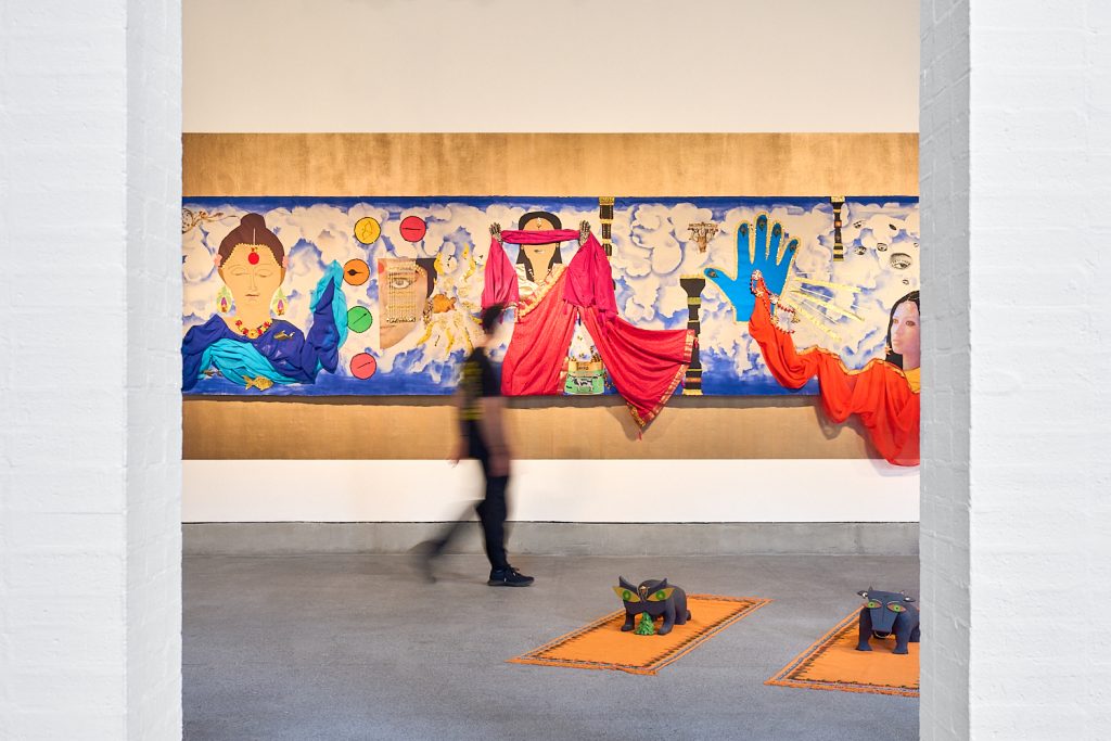 A long exposure shot of a gallery space. a blurred figure walks past a brightly coloured collage depicting women in Indian clothing. By the feet of the figure are two sculptures - one of a cat, one of a cow - made from clay.