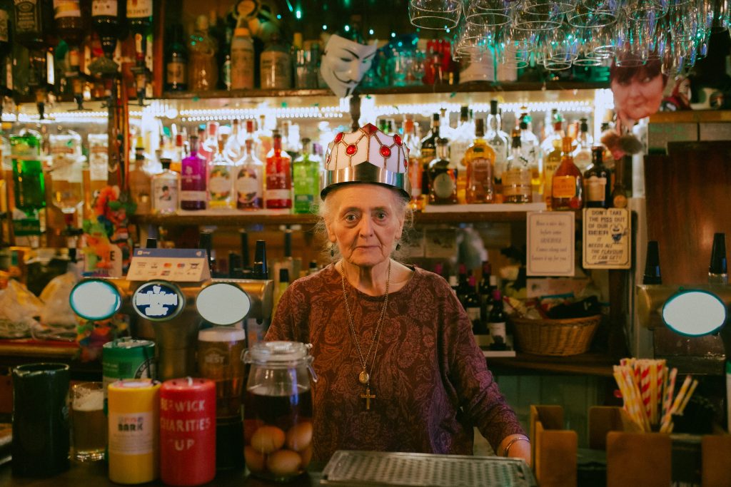 A woman stood behind a bar wears a crown made from metal and glass.