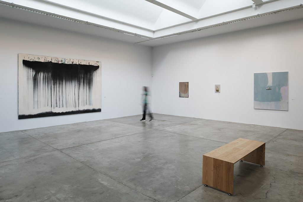 Big black paint streak tumbling down the otherwise blank canvas on the left, a blurred human figure moving through the room towards it
