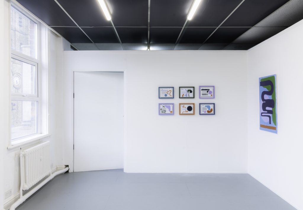 A white wall gallery with 6 small bright abstract paintings arranged into a rectangle on one wall