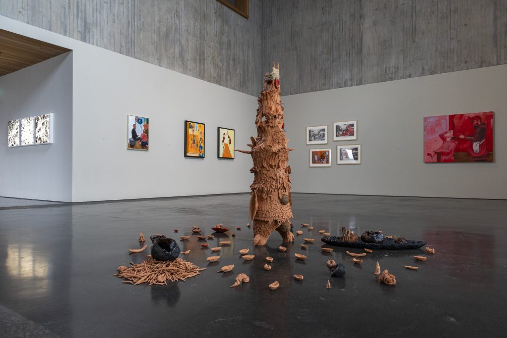 A fractured clay sculpture scattered on the floor sits in the middle of a concrete gallery space, behind it, on the walls, paintings and photographs