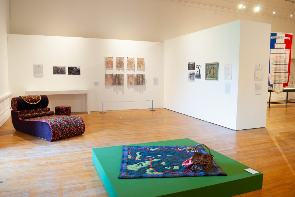 A big gallery with a dark chaise longue in the back corner and in the foreground a textured picnic blanket on a low green plinth with a picnic basket