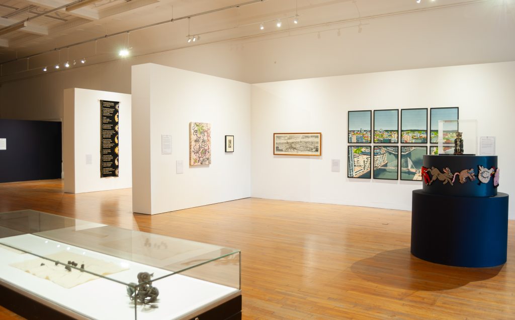 A bright white gallery with a blue cylindrical sculpture in the middle and framed prints on the walls