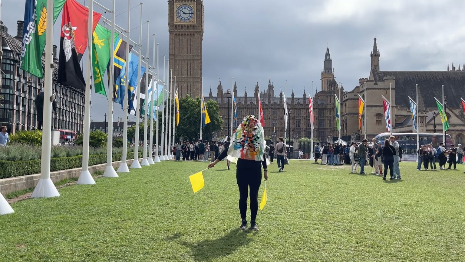 A person with multicoloured crinkle cut paper covering their face stands in front of Westminster Abbey with their arms by their sides, pointing yellow flags in each hand