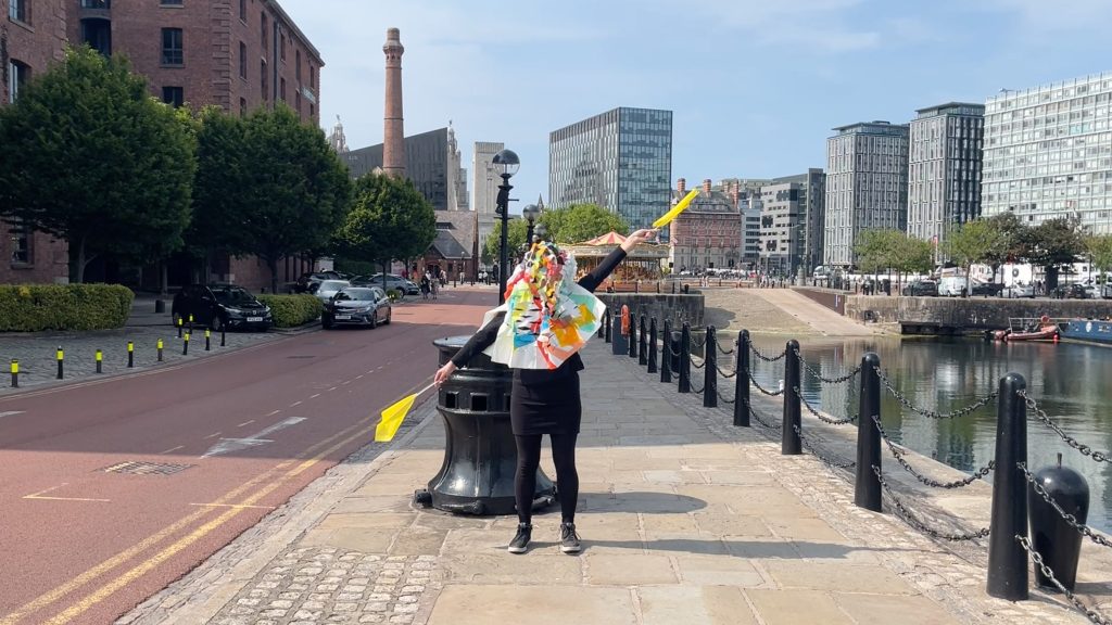 A person with multicoloured crinkle cut paper covering their face stands by a quay in Liverpool city centre with their arms wide, pointing yellow flags in each hand