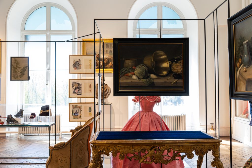 Photograph of an art gallery interior showcasing framed paintings, sketches, and a vintage gramophone displayed within glass cases. Central focus on a pink Victorian-style dress behind a glass case with ornate wooden table in foreground, highlighting historical and artistic exhibits.