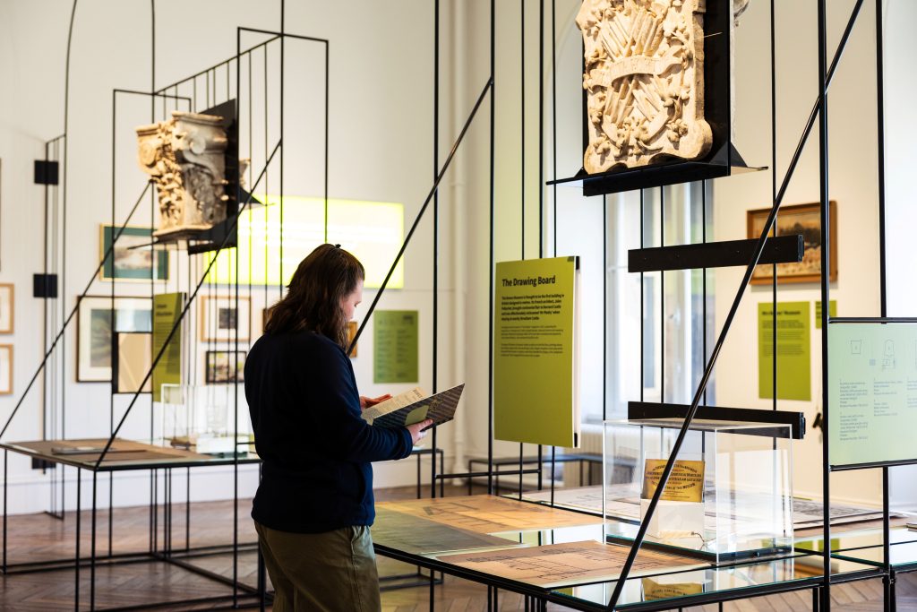Photograph of a museum exhibition room featuring historical artifacts displayed on suspended metal frames with informational panels in green and yellow. A person is reading a booklet while examining detailed stone carvings and documents, highlighting educational and cultural engagement.