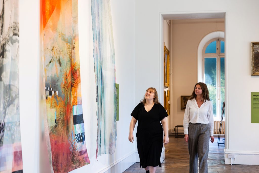 Photograph of two women walking through an art gallery with large, colourful abstract paintings hanging on white walls. The scene highlights natural light from tall windows, wooden flooring, and framed artworks, creating a calm and contemplative atmosphere.Photograph of two women walking through an art gallery with large, colourful abstract paintings hanging on white walls. The scene highlights natural light from tall windows, wooden flooring, and framed artworks, creating a calm and contemplative atmosphere.
