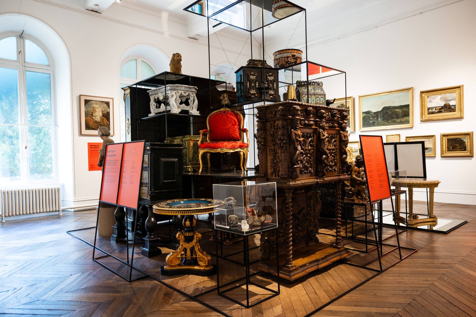 Photograph of a museum exhibit showcasing intricately carved antique furniture and decorative objects arranged on black metal stands with red information panels. The room features large arched windows, wooden flooring, and framed paintings on white walls, highlighting historical craftsmanship and artistic heritage.