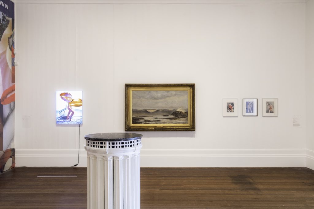 A lightbox image of a photo montage including a woman standing before a boat, hung beside a historical brown landscape painting