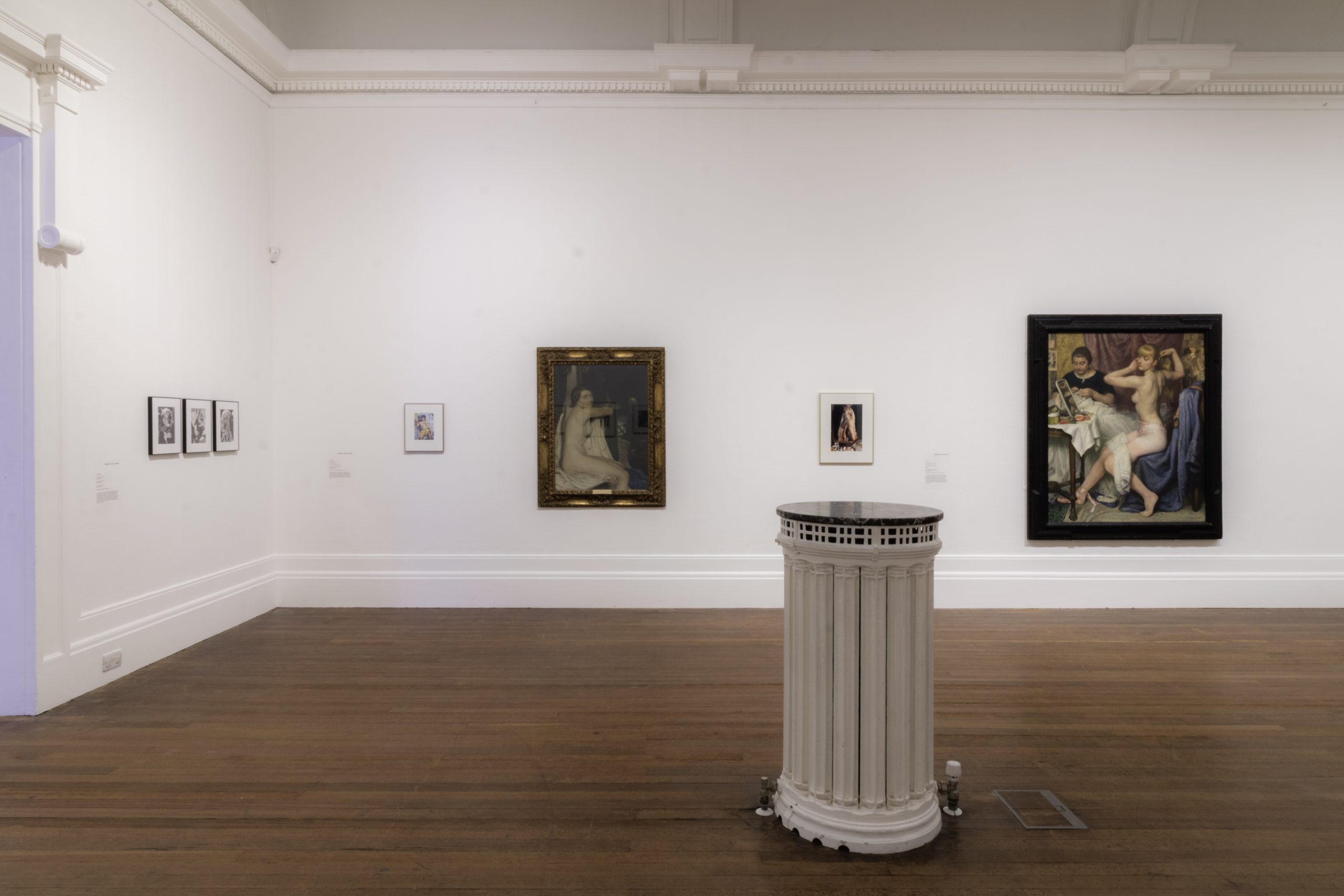 A white gallery with two large traditional nudes interspersed with smaller artworks