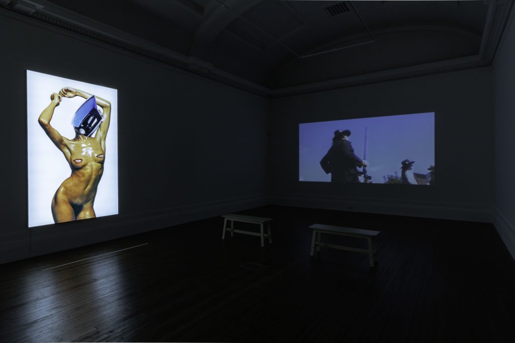 A bright image of a naked body with the face replaced by a metallic item projected into a dark room