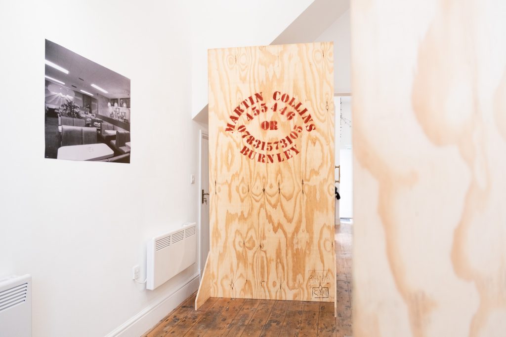 A black and white photo of a bar or hotel lobby on the left, straight ahead is a big sheet of unfinished wood printed with the words MARTIN COLLINS BURNLEY and a phone number