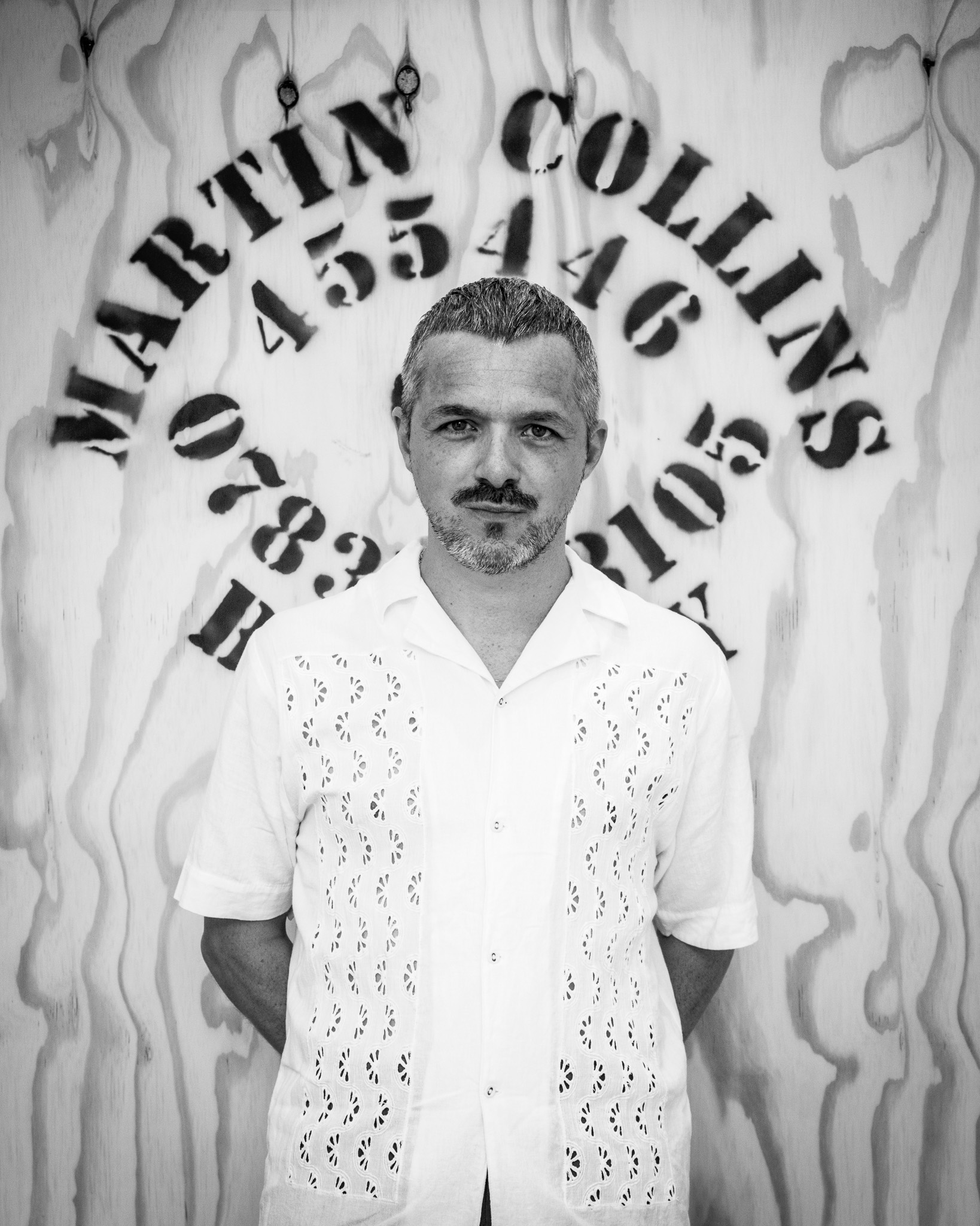 Flanagan stands in front of the MARTIN COLLINS sign in black and white, he has light skin and dark hair with trimmed facial hair and wears a white shirt