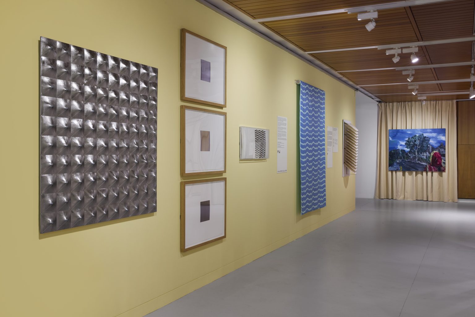 A yellow wall with many works on paper hung along it, including a big silver square, some smaller works in frames and a long blue print