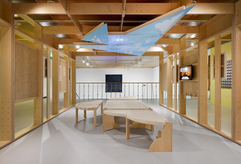 A wide view of the gallery with Disshou's manji in the middle of a built wooden structure with open walls and a room, and the blue painting above like the sky