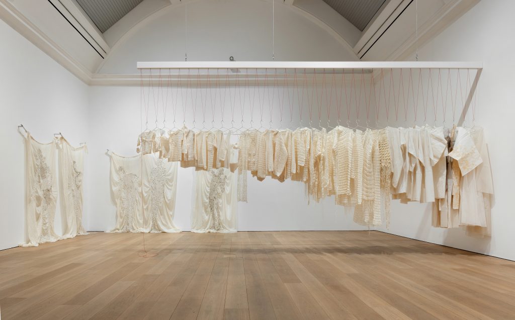 A white room filled with white fabrics, on the back wall some tapestries and in the centre a clothes rail seen side on, with shite clothing ordered in size, tiny and short on the left growing longer towards the ground on the right
