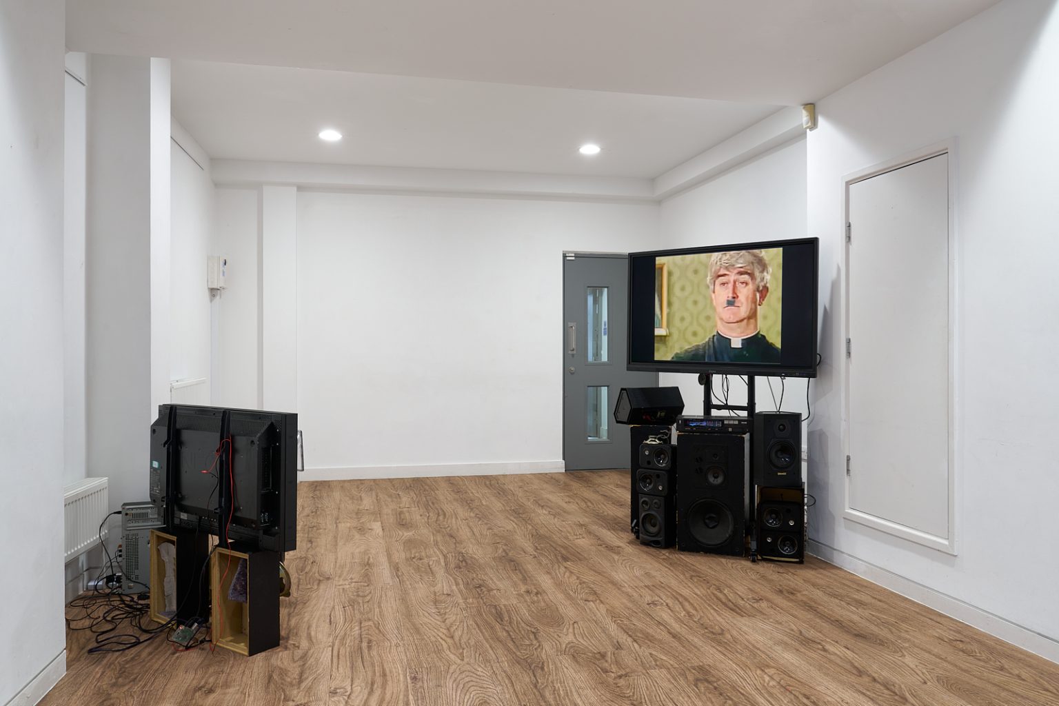 Two flatscreen monitors face each other in a white gallery space. Each sits atop a pile of various speakers, wires trail around. One screen isn't visible, the other shows a still from TV series 'Father Ted'.