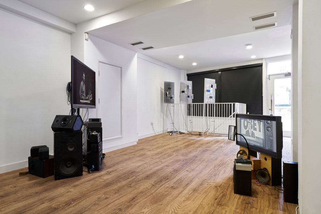 A white gallery space with wooden flooring. In the foreground are two flatscreen monitors facing each other and sitting on top of some broken or deconstructed analogue speakers. In the background are three metal panels with smartphones attached to them.
